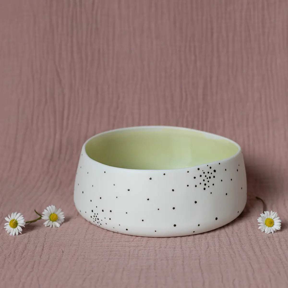 The Breda Bowl, dotty from the Urchic Porcelain collection, featuring a white porcelain bowl adorned with small black dots on its exterior, sits gracefully on a textured pink surface, accompanied by three delicate white flowers.