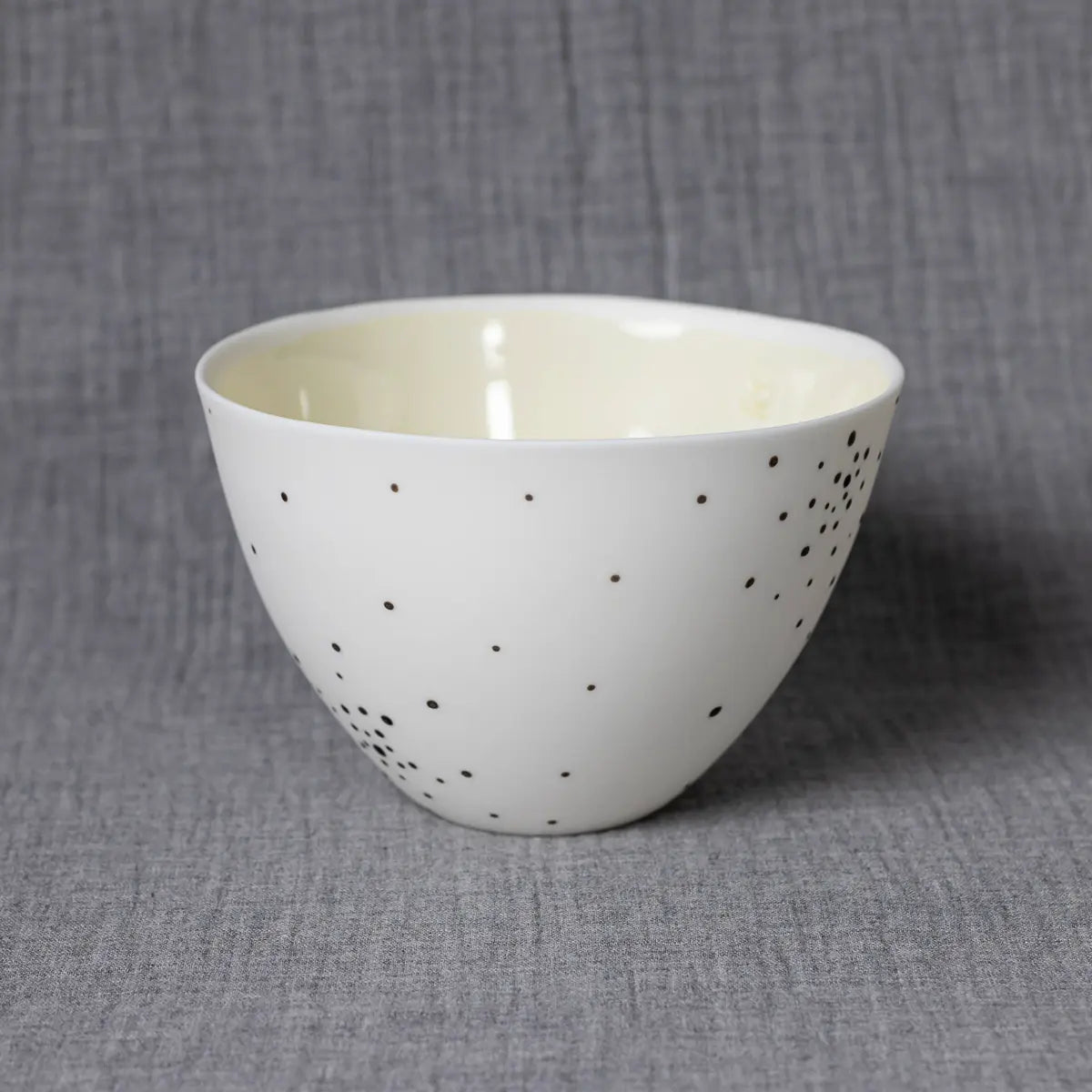 The Martinka Bowl, dotty by Urchic Porcelain is a white ceramic bowl with small black polka dots, set against a gray textured background. This versatile piece can enhance any dining setting and adds a charming decorative element to your table.