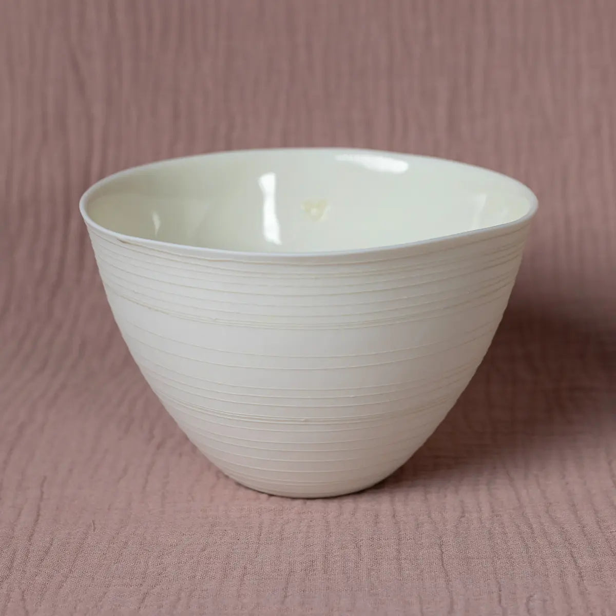 The Martinka Bowl with lines from Urchic Porcelain is a versatile piece of dinnerware, showcasing a subtle ridged texture on plain, off-white ceramic. It is elegantly set against a pink, textured fabric background.