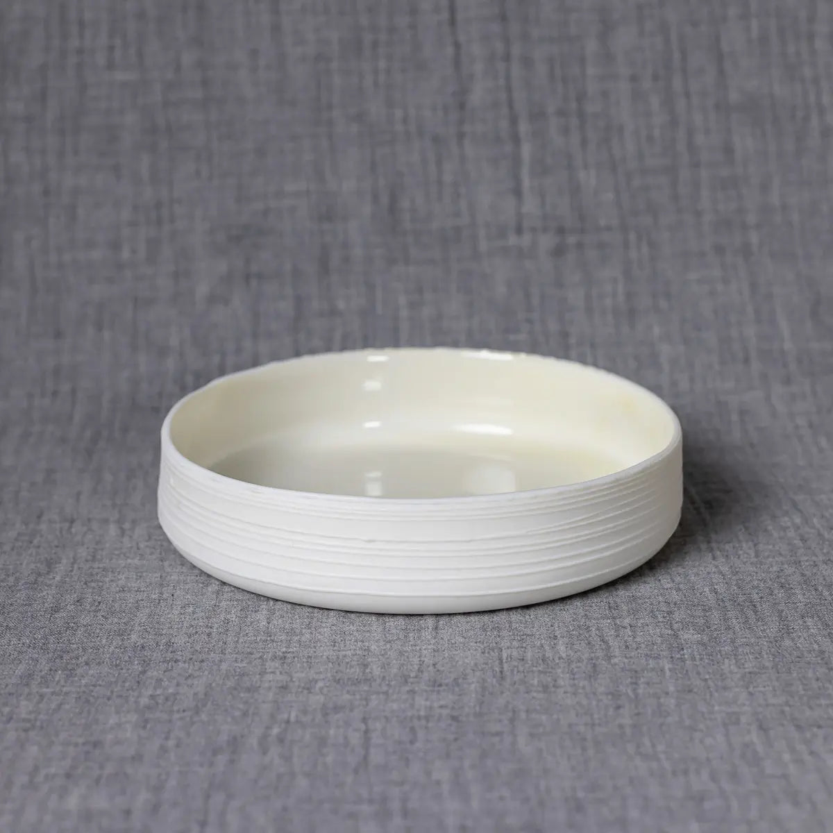 The Breda Mini Bowl with lines from Urchic Porcelain, featuring horizontal ridges on its outer surface, is set against a gray fabric background. This shallow bowl effortlessly blends into any setting, exuding charm and sophistication.