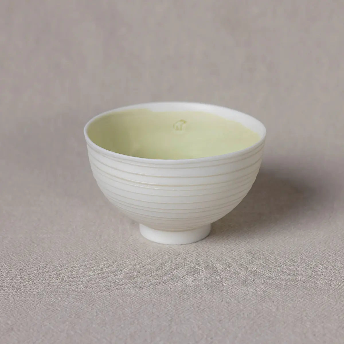 The Vida Bowl by Urchic Porcelain, a white, ribbed ceramic piece showcasing timeless elegance, is filled with a light yellow substance and set against a beige textured background.