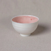 A small, white Vida Bowl from Urchic Porcelain with subtle horizontal lines on the exterior and a pink interior sits on a textured beige surface, exuding timeless elegance.