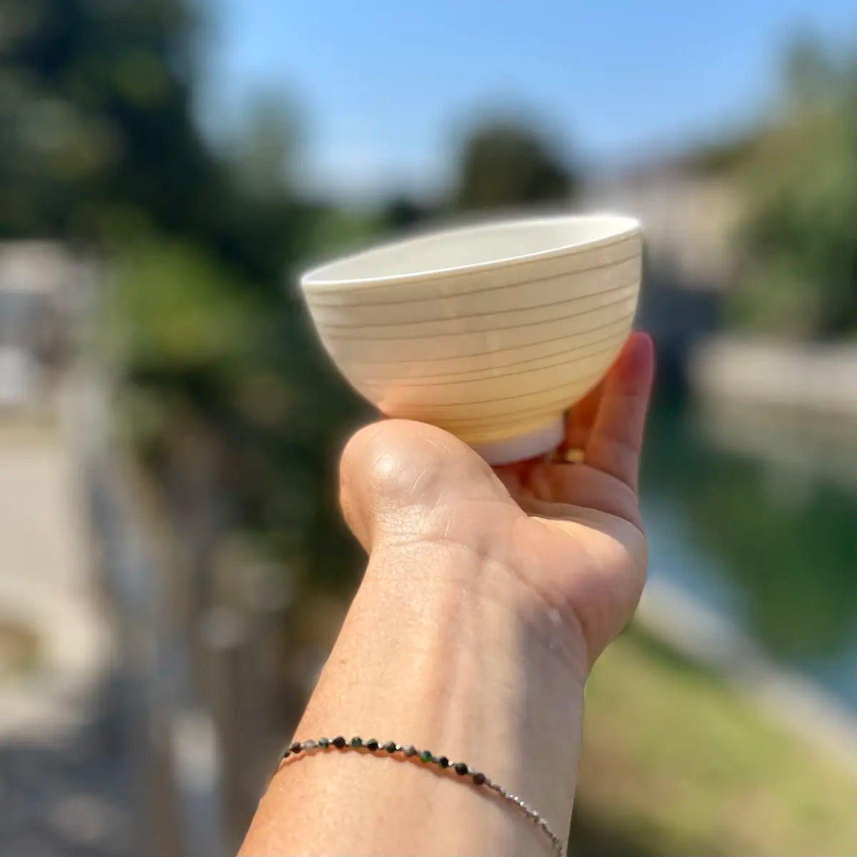 A hand adorned with a bracelet holds up an Urchic Porcelain Vida Bowl outdoors, showcasing its timeless elegance against a blurred backdrop of trees and a serene body of water.