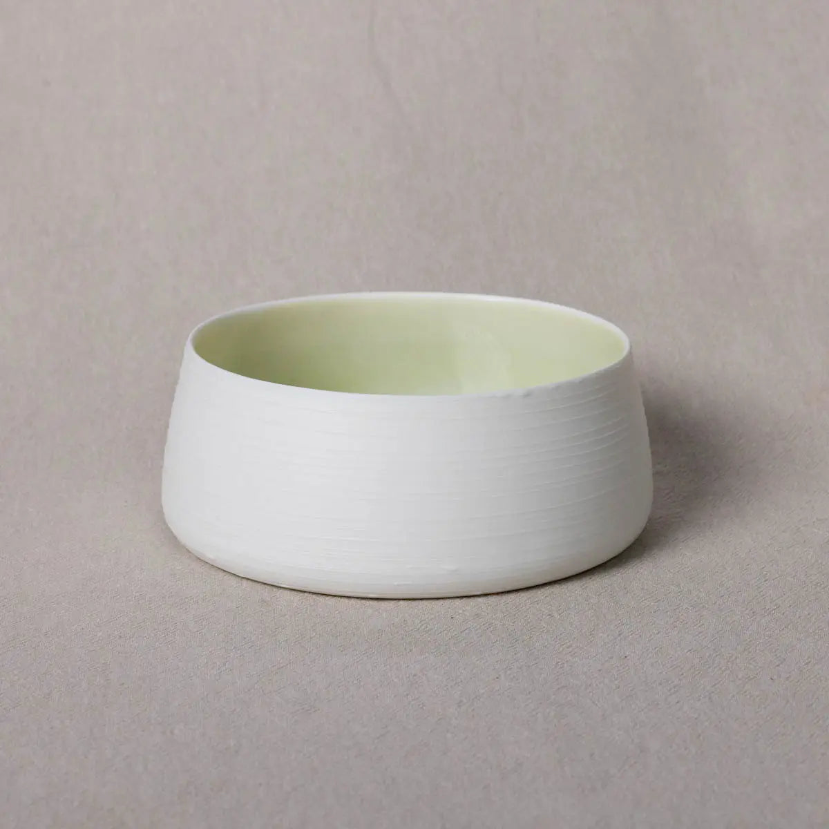 A white, round Breda Bowl with lines from Urchic Porcelain featuring a smooth, slightly tapered exterior and a light yellow interior, set against a neutral background.