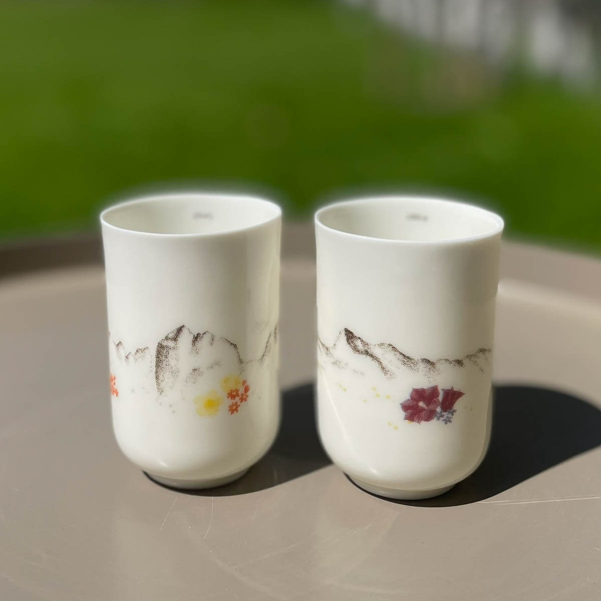  Peak Peace from Urchic Porcelain feature white ceramic with elegant mountain and flower patterns inspired by Slovenian mountain ranges. They sit on a round surface, with green grass softly blurred in the background.