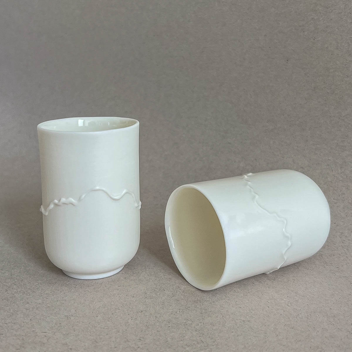 Two white ceramic cups with uneven textures and wavy patterns are displayed, one standing upright and the other lying on its side, on a neutral background. This handmade Soulmates Cup Set of Two Cups, Alpine Poetry by Urchic Porcelain, MADE IN SLOVENIA, evokes the rugged beauty of Slovenian mountain ranges.