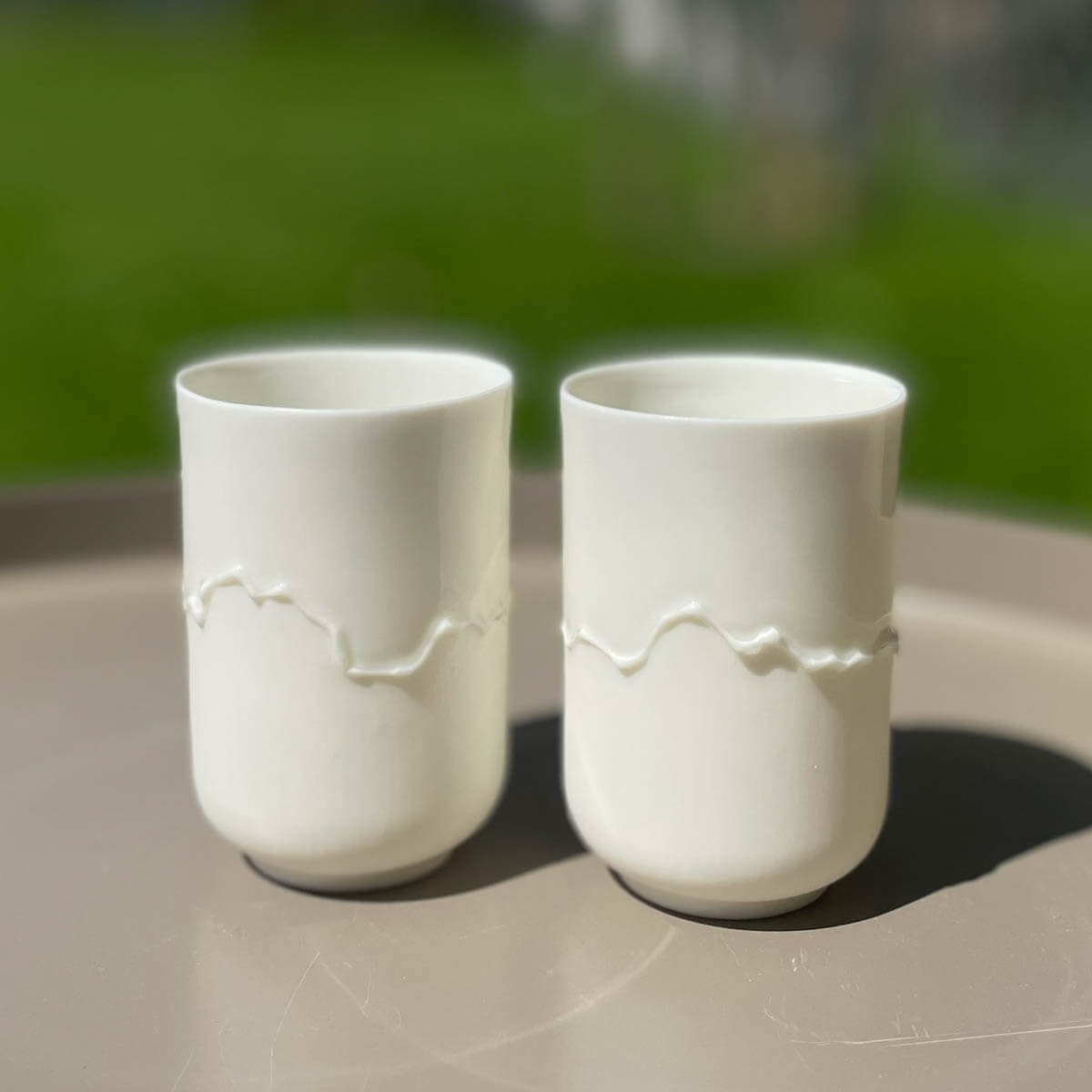 The Soulmates Cup Set of Two Cups, Alpine Poetry by Urchic Porcelain are displayed on a beige surface with a blurred green background, proudly showcasing the artisanal charm of a handmade porcelain coffee set inspired by Slovenian mountain ranges. These white ceramic cups feature a wavy raised pattern in the middle that enhances their elegant design.