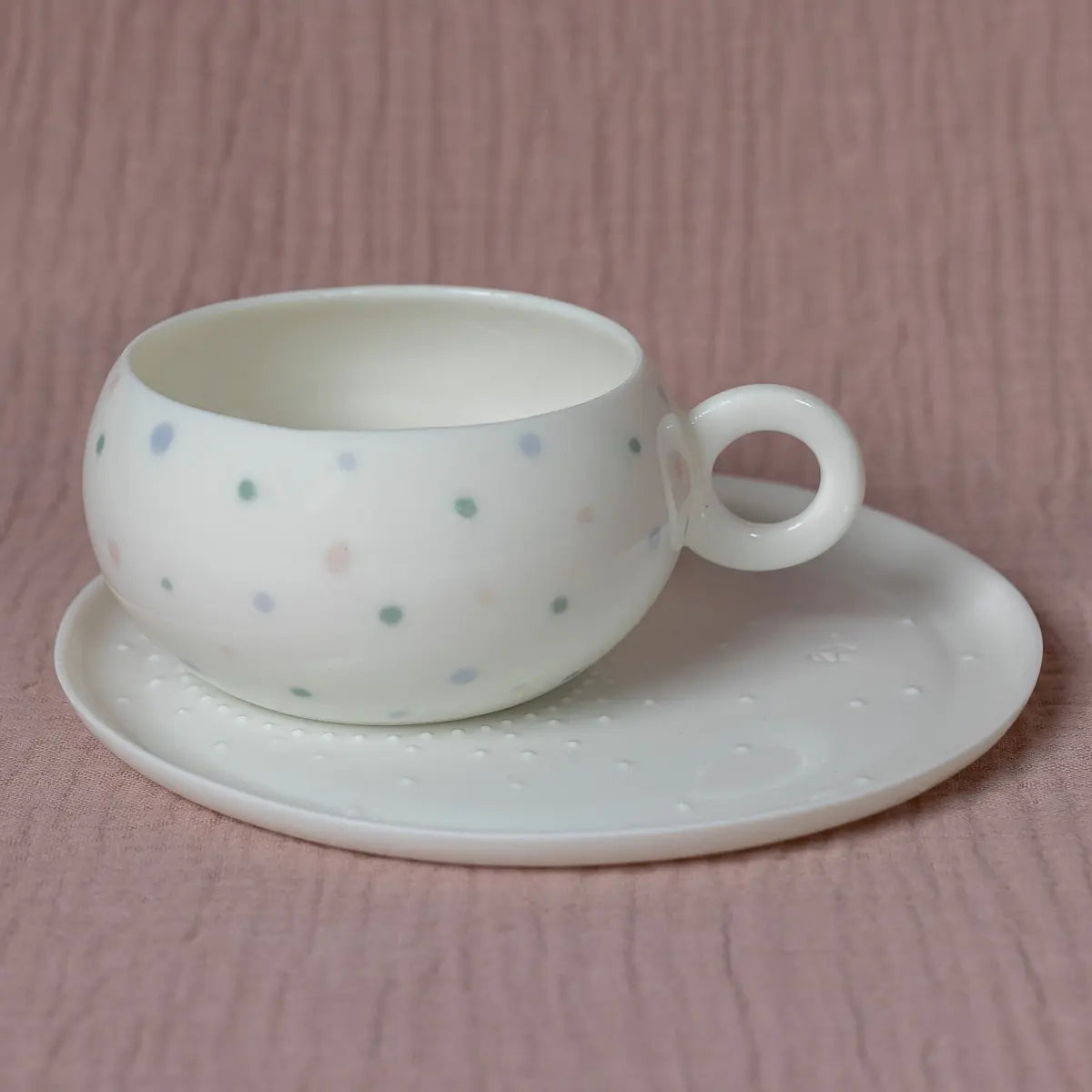 A Dama Tea & Coffee set by Urchic Porcelain, featuring a white teacup adorned with pastel polka dots, rests elegantly on its matching white saucer. The backdrop is a textured pink fabric, creating an ideal setting for enjoying a soothing cup of green tea.