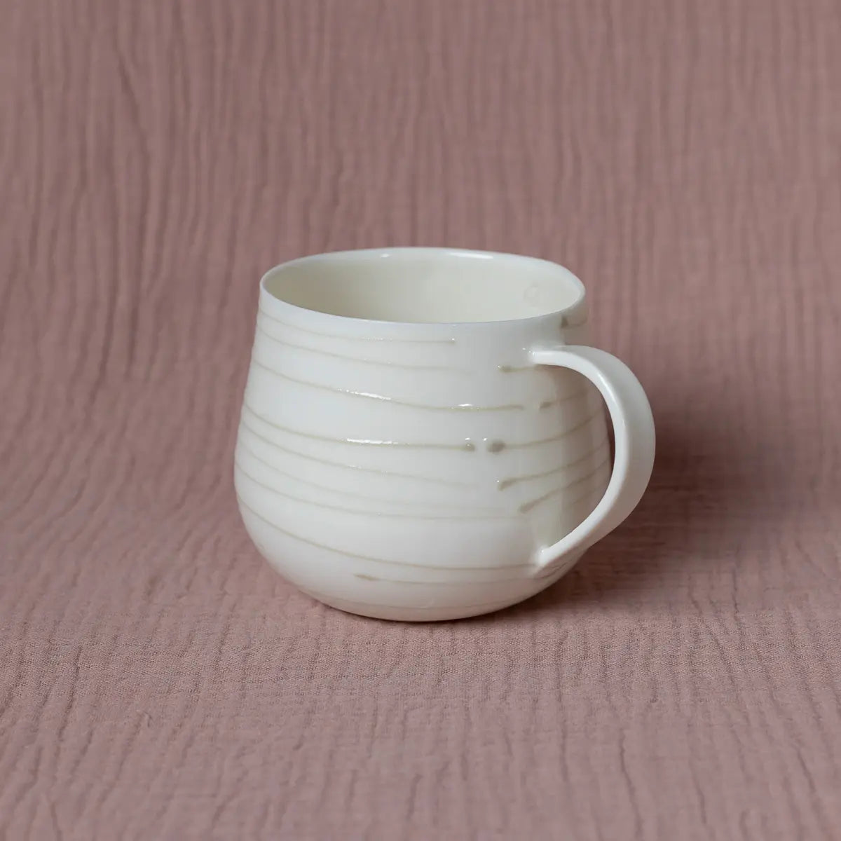 A white Nova Cup with smooth 3D lines, from the Urchic Porcelain brand, is placed against a pink fabric-like background. The cup, perfect for savoring herbal tea, boasts a rounded shape and large capacity with an easy-to-hold handle.