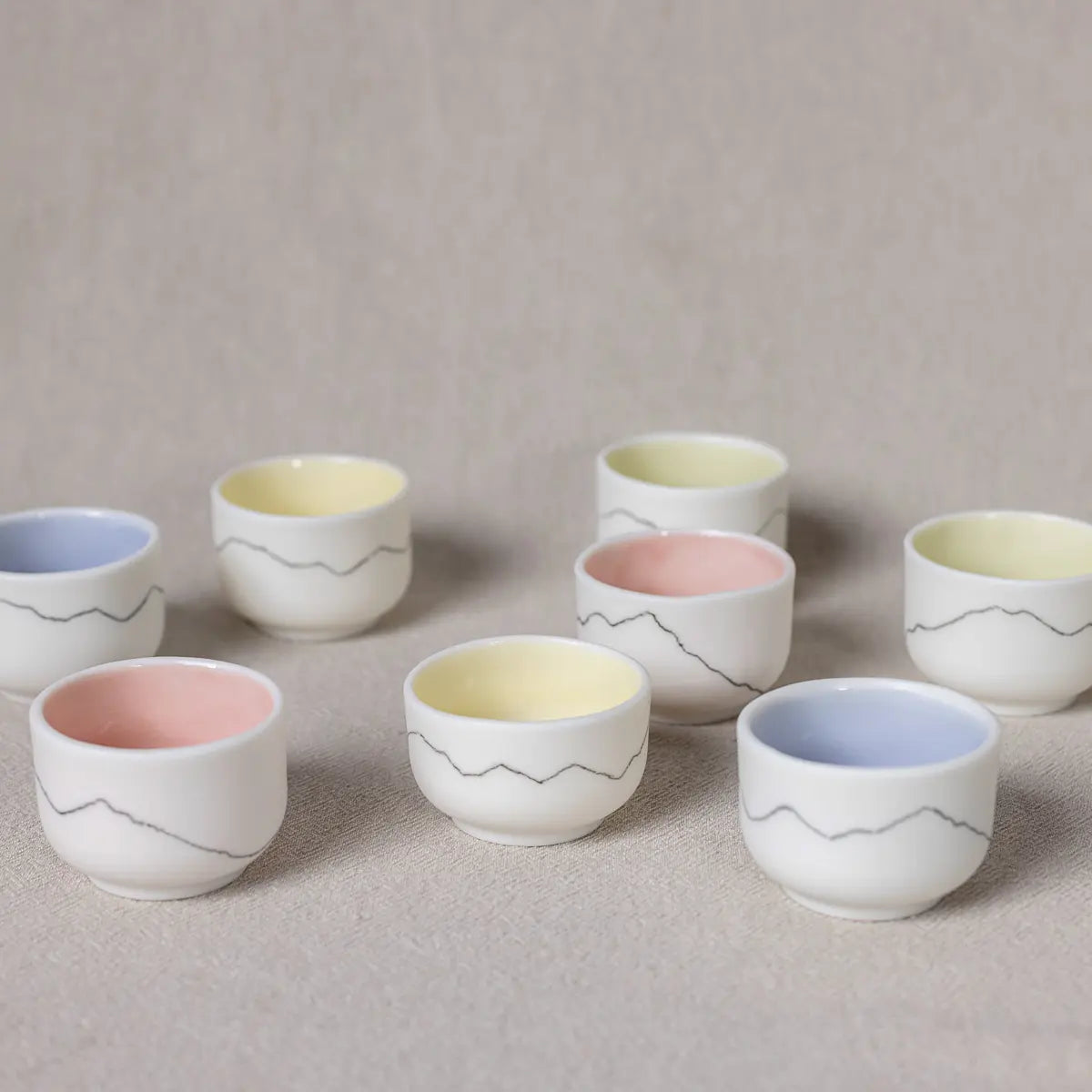 A set of Kiki mini ceramic cups by Urchic Porcelain, featuring four vibrant mountain designs and pastel interiors in pink, yellow, and blue, beautifully arranged on a beige surface.