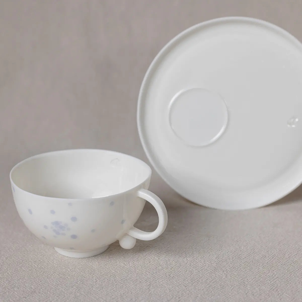 The Snow White Cup by Urchic Porcelain, a white ceramic teacup adorned with light blue spots and part of a handcrafted porcelain tea set, is placed next to an overturned white saucer on a beige fabric surface.