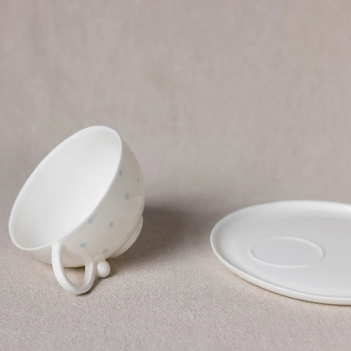 A handcrafted Urchic Porcelain Snow White Cup adorned with polka dots rests on its side next to an empty white saucer, both set against a neutral background. This elegant tea set features an ergonomic design.