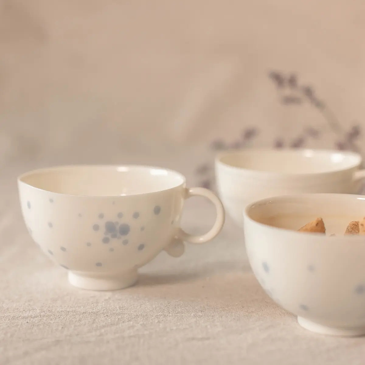 Three Snow White Cups by Urchic Porcelain, featuring blue speckles, are arranged on a light-colored surface. One handcrafted cup has two small pieces of biscuit.