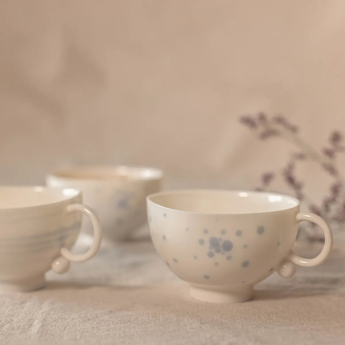 Three Snow White Cups from Urchic Porcelain, adorned with subtle blue patterns, grace a beige fabric surface, showcasing the elegance of this handcrafted porcelain tea set. In the background, a blurred purple plant adds to the serene ambiance.