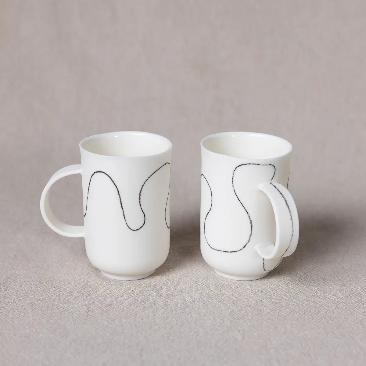 Two Urchic Porcelain Soulmates coffee cups, each adorned with a soft waves pattern, sit on a gray surface. The handles are to the right, and the background is a textured neutral gray, presenting an ideal setting for your morning brew.