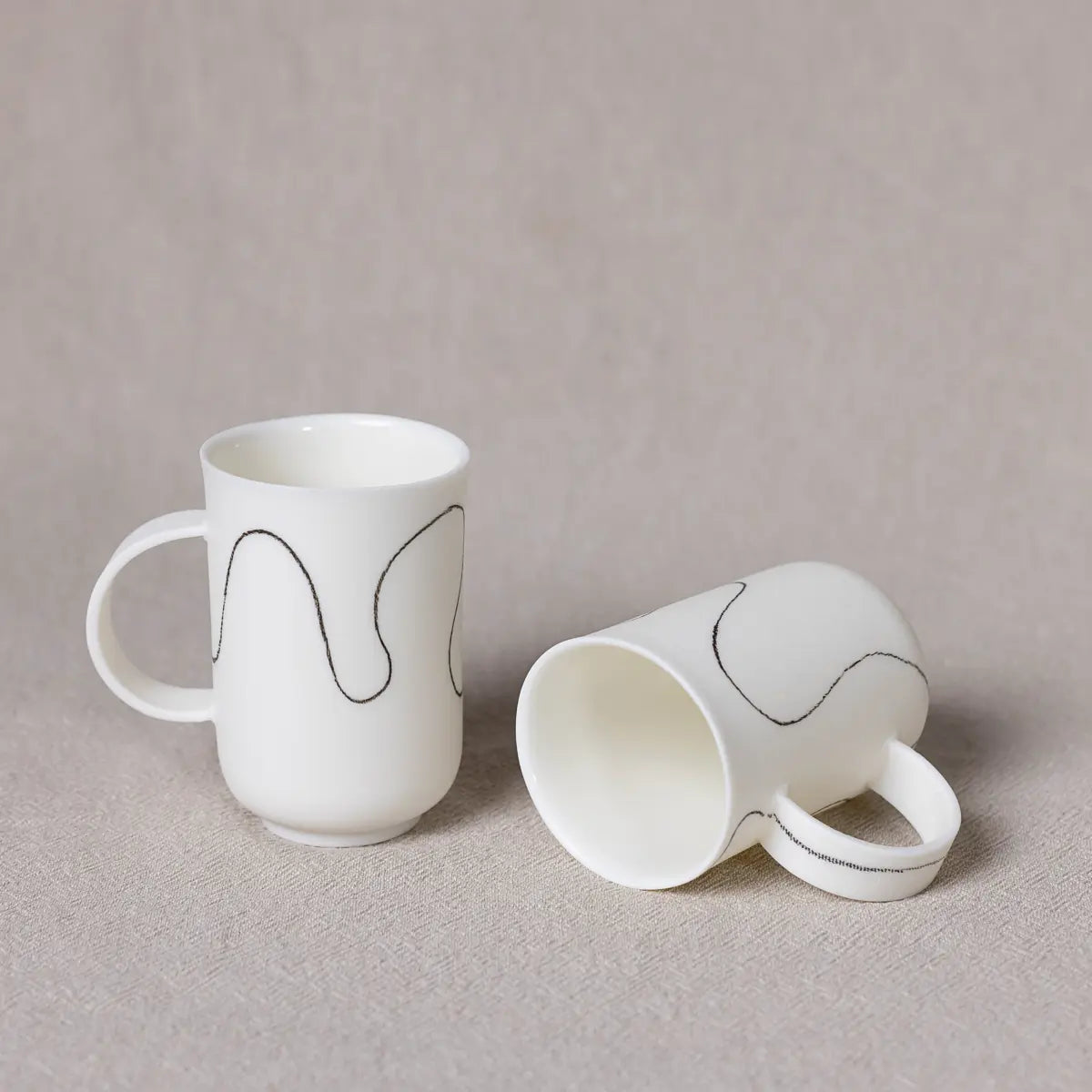 Two minimalist coffee cups from Urchic Porcelain's Soulmates porcelain coffee set, each featuring soft waves patterns in black lines on a white background; one cup is upright while the other lies on its side, both displayed on a neutral fabric backdrop. Ideal as a soulmates set.