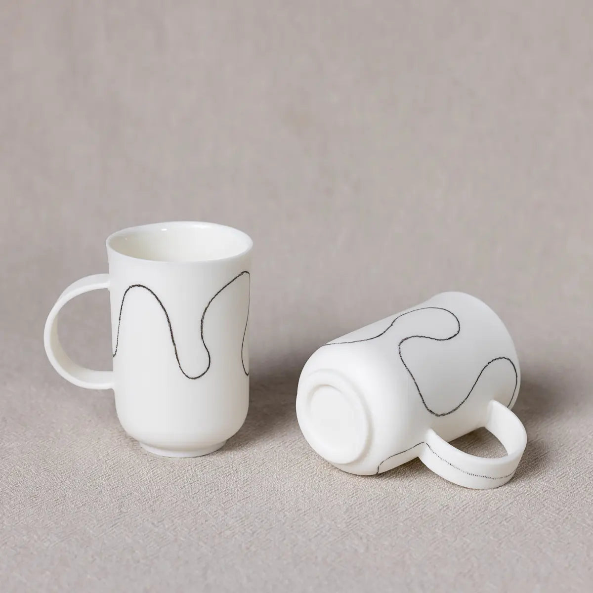 Two white handmade porcelain cups with soft wave patterns, one standing upright and the other lying on its side, are displayed on a light beige surface. This Soulmates porcelain coffee set of two cups by Urchic Porcelain is perfect for coffee lovers who appreciate unique artistry in their everyday rituals.