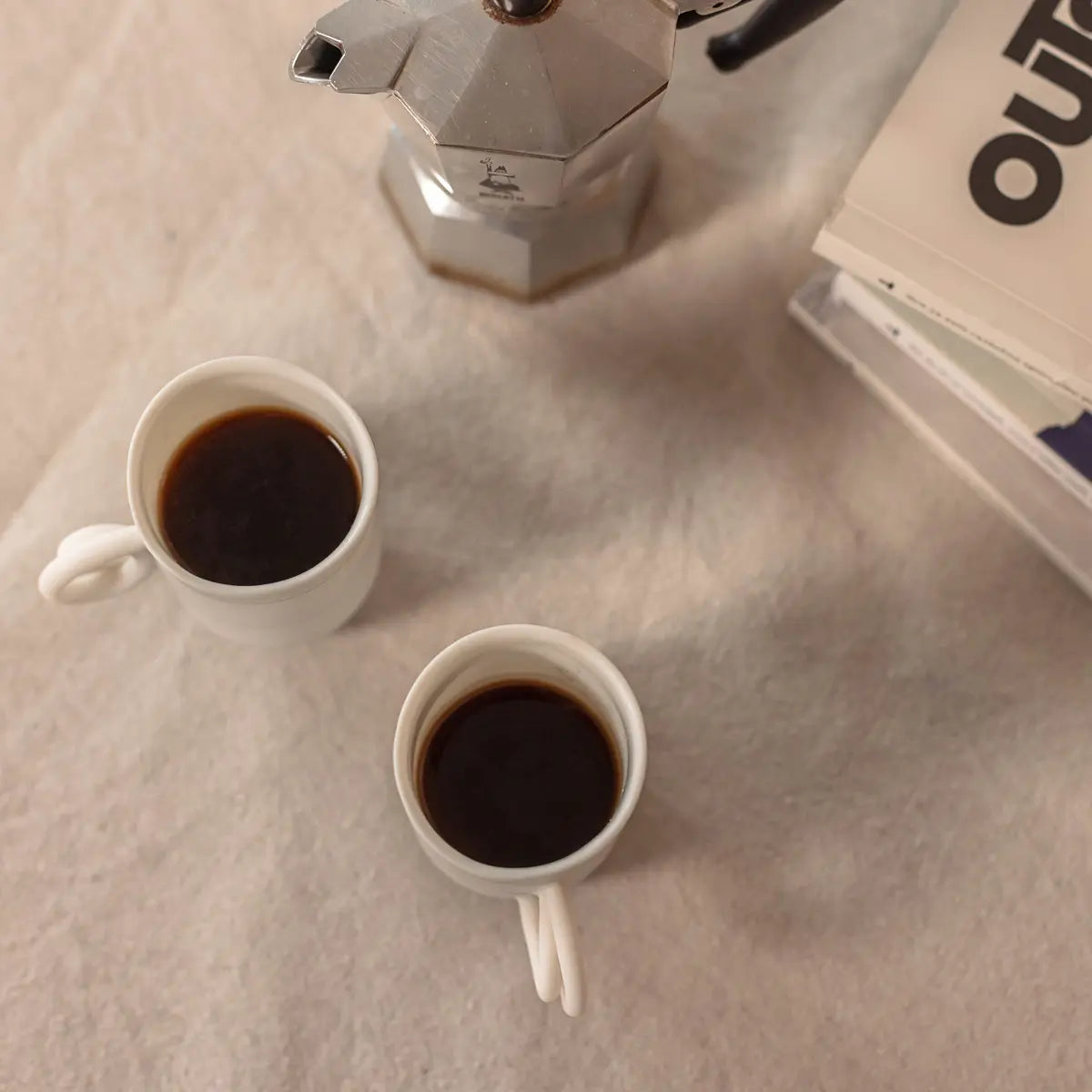 Two handcrafted mugs from the Soulmates porcelain coffee set of two cups, featuring a delicate Lace pattern by Urchic Porcelain, are filled with black coffee and placed on a table alongside a stovetop espresso maker and a stack of books.