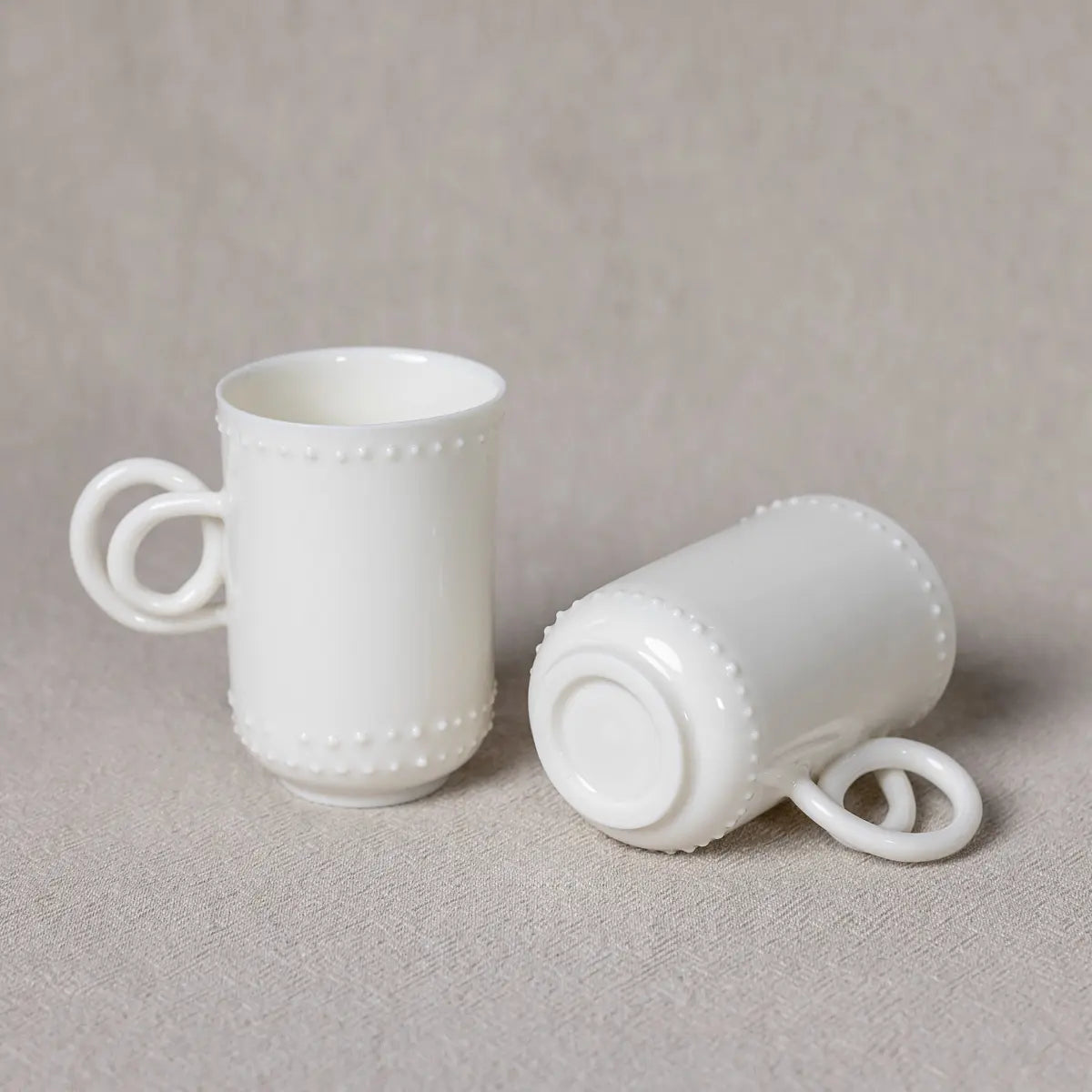 A pair of handmade cups from the Urchic Porcelain Soulmates coffee set, featuring a delicate Lace pattern. One cup stands upright while the other lies on its side on a beige surface, making an elegant display.