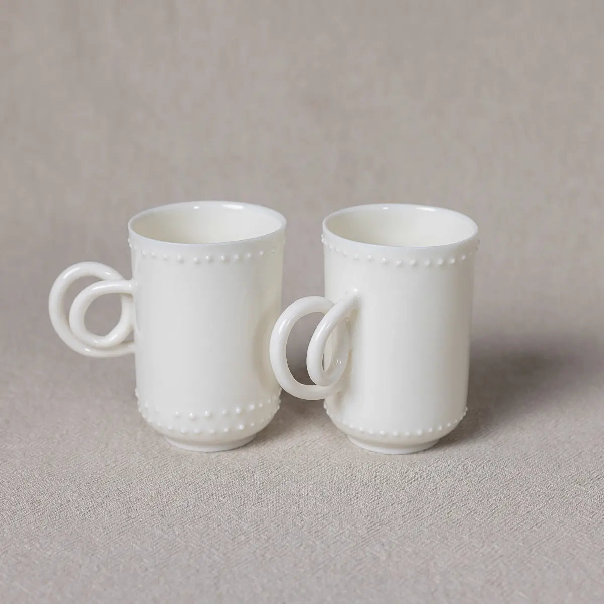 The Soulmates porcelain coffee set of two cups by Urchic Porcelain, featuring a delicate Lace pattern and twisted handles, is elegantly displayed on a neutral-colored surface, creating a charming arrangement.