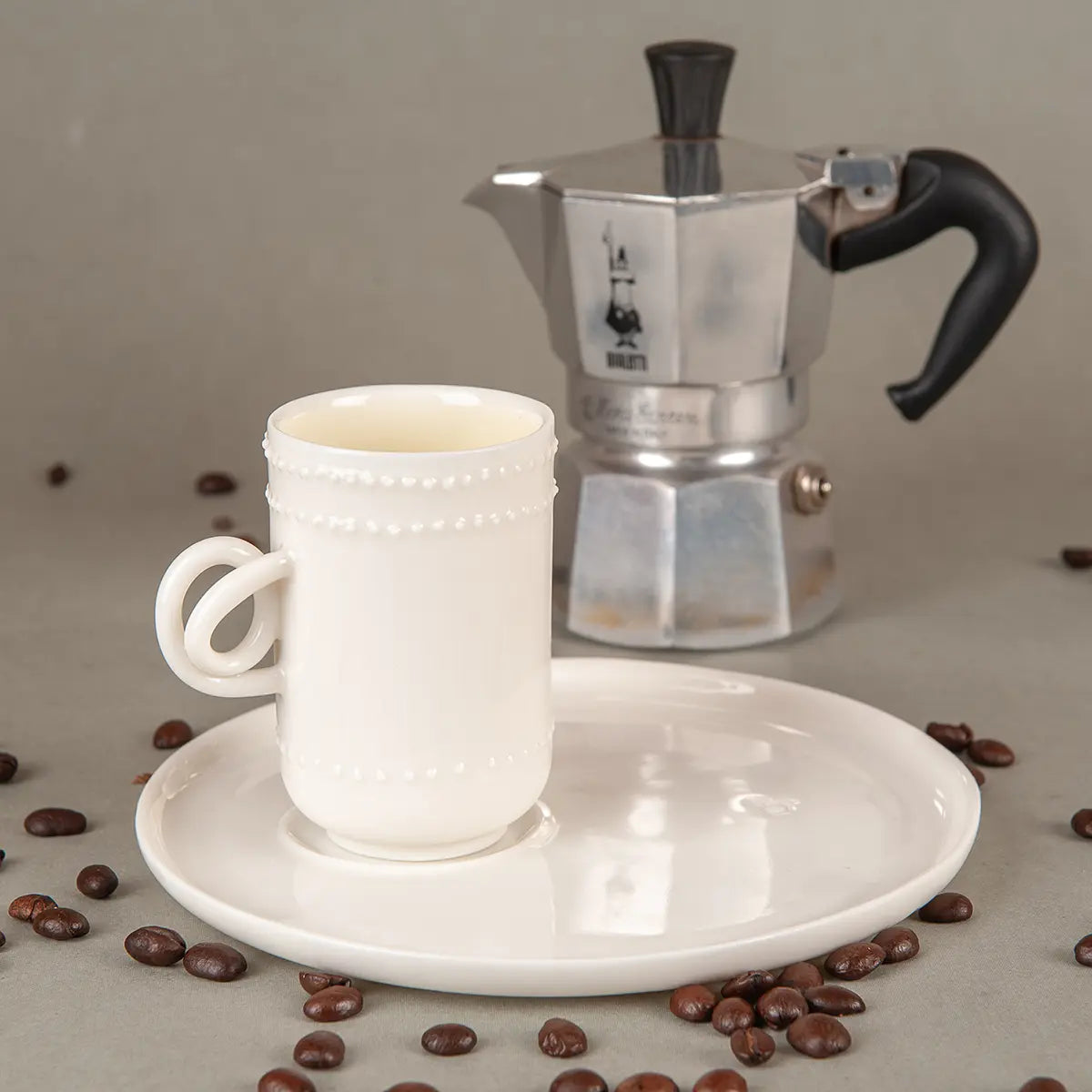 A Soulmates porcelain coffee cup from the elegant two-cup Lace pattern set by Urchic Porcelain, positioned on a matching saucer and surrounded by coffee beans, with a silver stovetop espresso maker in the background.