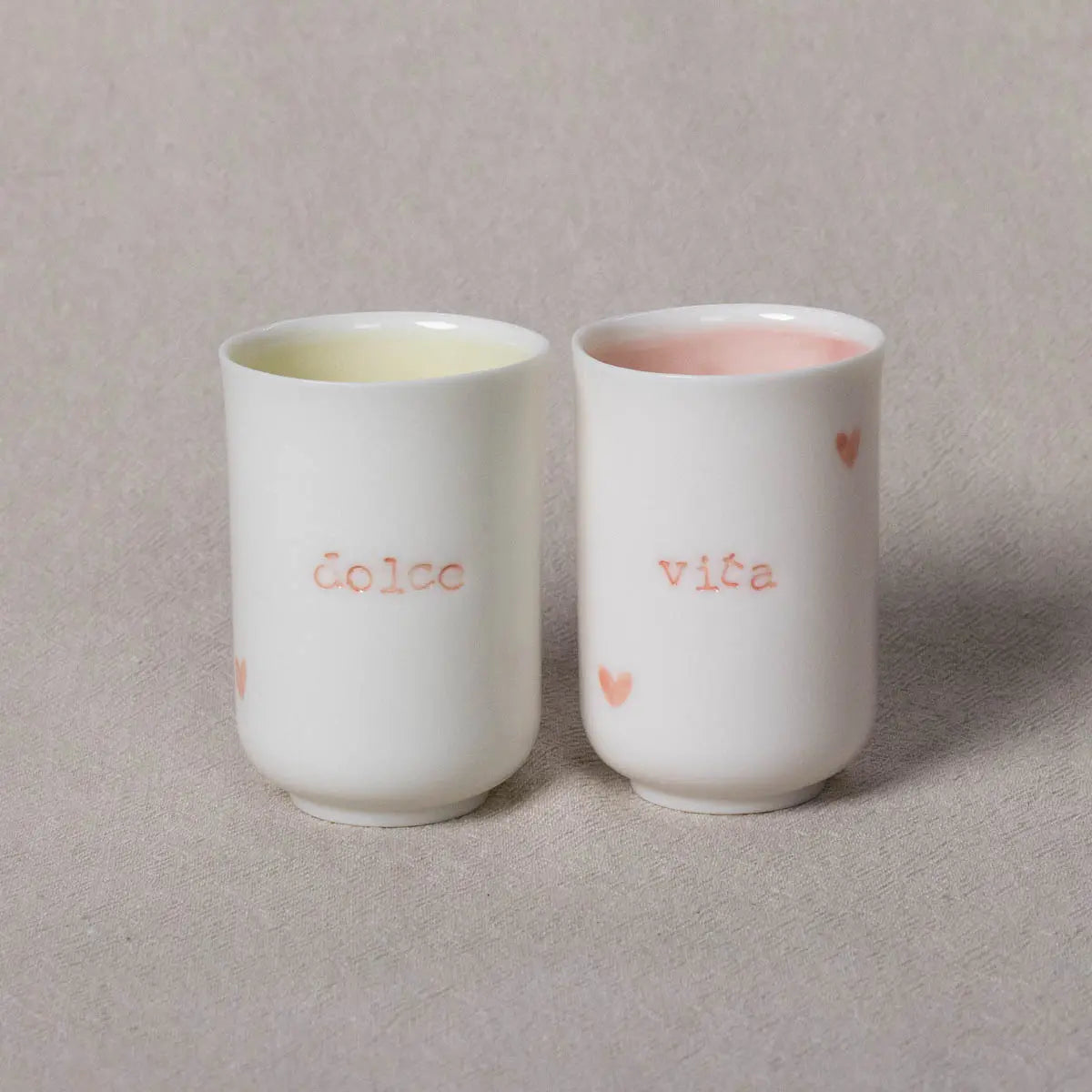 The Soulmates porcelain coffee set by Urchic Porcelain includes two white cups, each with a light pink and yellow interior. One cup features the "dolce" inscription, while the other showcases "vita," perfectly encapsulating Dolce Vita's essence. Small heart designs embellish each cup, all placed on a beige fabric surface—truly the ideal Soulmates set.