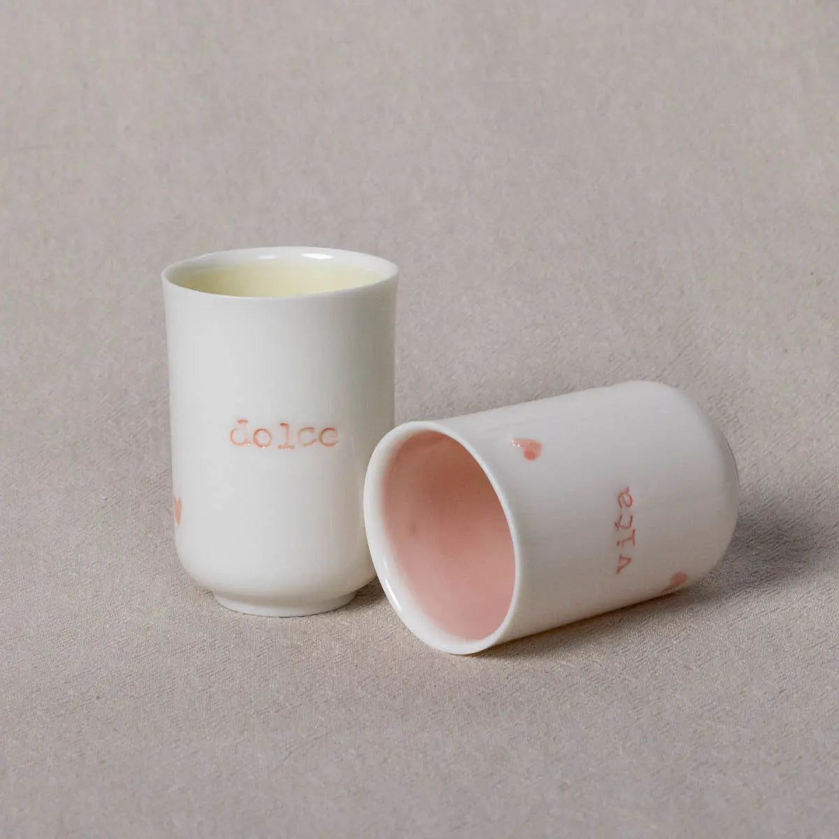 Two white ceramic cups from the Urchic Porcelain Soulmates set, one standing upright with the inscription "dolce" and the other lying on its side with the word "vita" and a pink interior. The background is a light beige fabric. This exquisite Soulmates porcelain coffee set perfectly captures the essence of Dolce Vita.