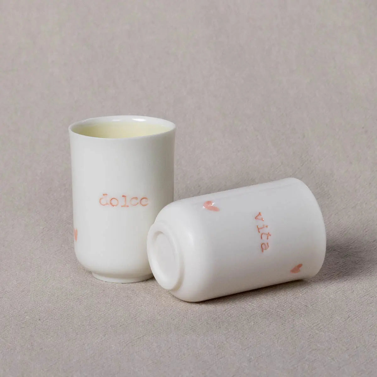 Two small white porcelain coffee cups from Urchic Porcelain's Soulmates collection, one standing upright and the other lying on its side. The standing cup has "dolce" written in pink, and the lying cup has "vita" written in pink. This Soulmates set perfectly captures the Dolce Vita vibe.