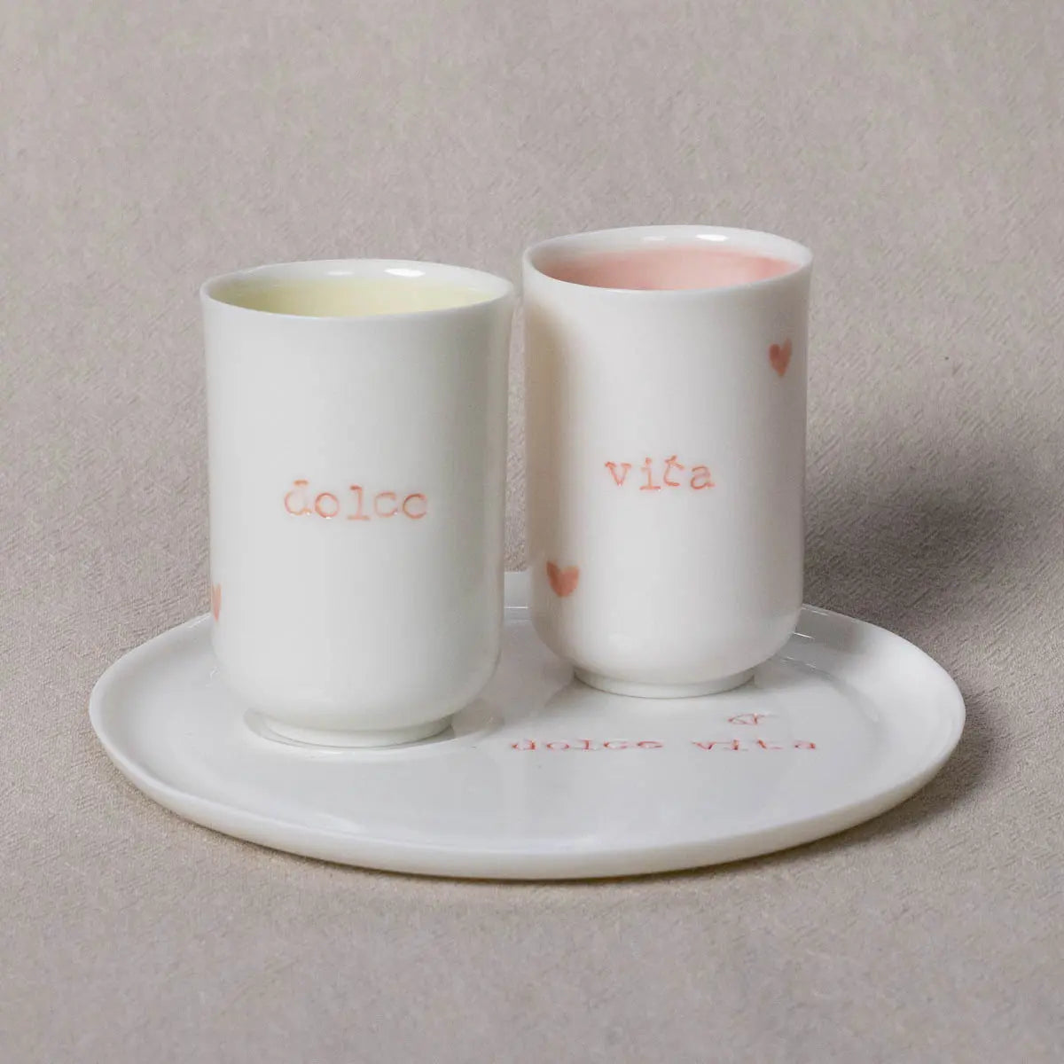 Two white porcelain coffee cups with the pink and gold inscription "Dolce Vita" from the Soulmates set by Urchic Porcelain stand on matching saucers. The cups feature small heart designs and are placed against a neutral background.