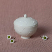 The Urchic Porcelain Sugar Bowl Vida is centered on a pink textured surface, accompanied by three small white daisies with green centers.