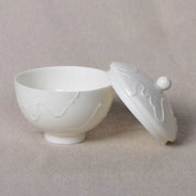 A white ceramic Urchic Porcelain Sugar Bowl Vida, with a lid placed beside it, featuring a subtle wavy pattern reminiscent of traditional porcelain sugar bowls, set against a beige backdrop.