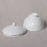 Two white ceramic pieces from Urchic Porcelain, the Sugar Bowl Vida with a spherical handle and a round lid, and the other resembling an upside-down bowl with a small circular opening on top, both featuring a subtle wave pattern. The versatile jar boasts an elegant design, while the porcelain sugar bowl fits comfortably in your palm.