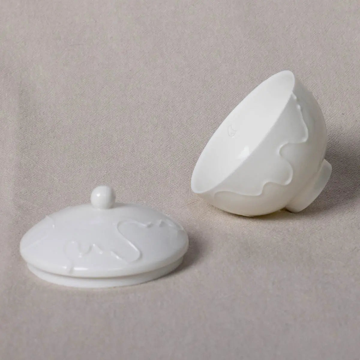 The Sugar Bowl Vida by Urchic Porcelain, a white porcelain sugar bowl adorned with a wavy pattern, is displayed on its side next to its matching lid featuring a small spherical handle, both resting on a light beige surface.