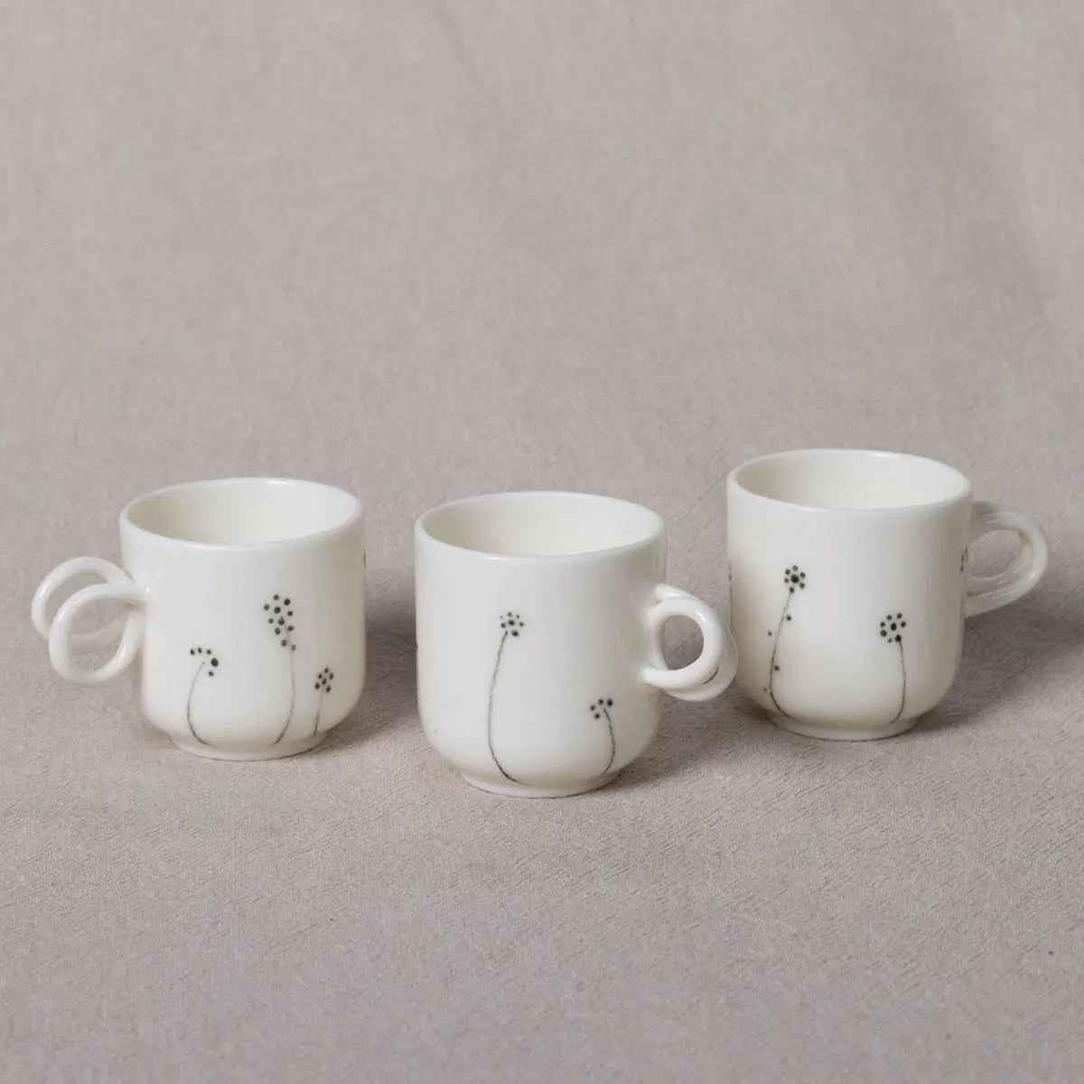 Three Urchic Porcelain Espresso Mini Cups, adorned with black floral designs and featuring twisted handles, are neatly arranged in a row on a beige fabric background.