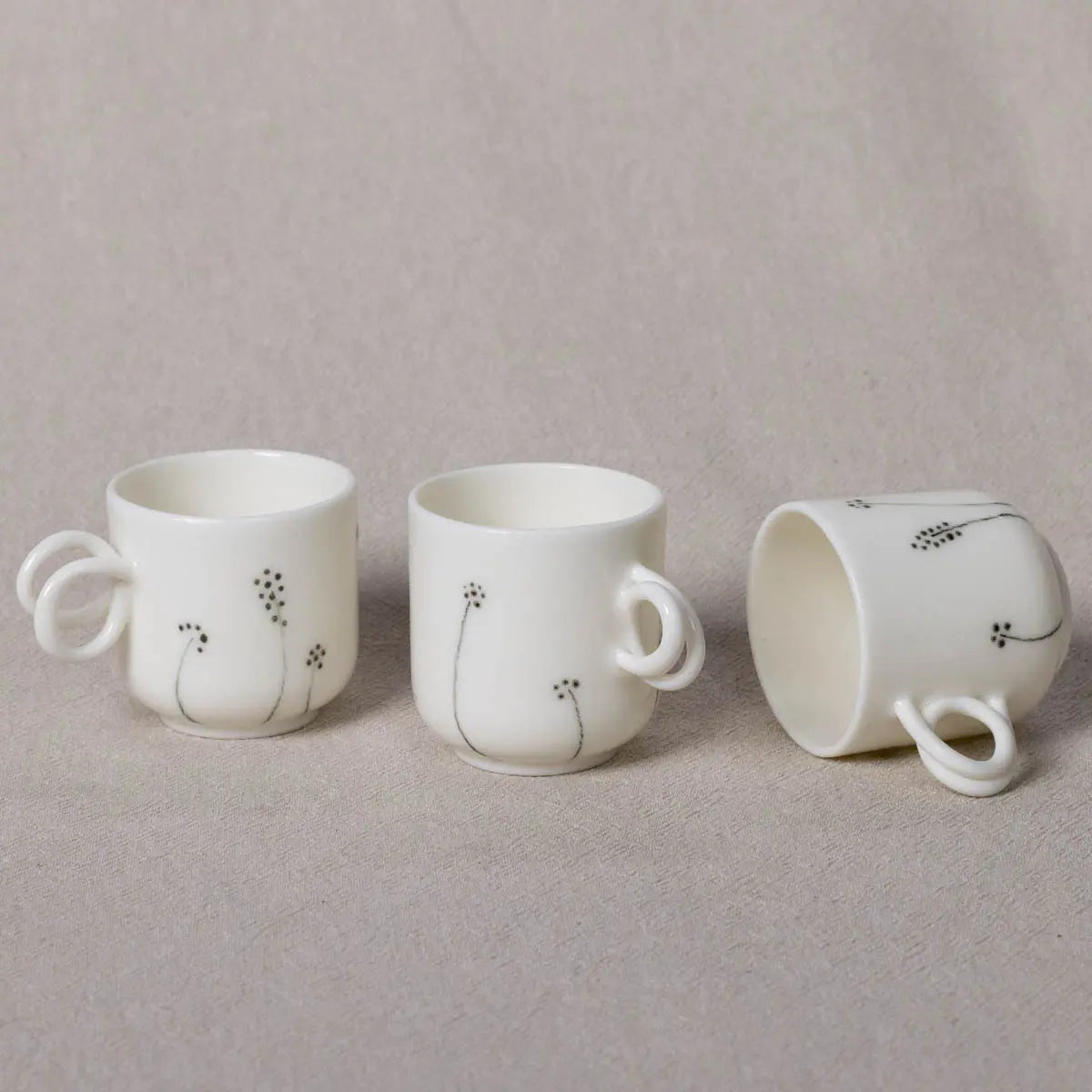 Three "Espresso Mini Cups, Meadow Flowers" by Urchic Porcelain, featuring twisted handles and decorated with a minimalist black floral design, are displayed. One hand-painted cup lies on its side on a beige surface.