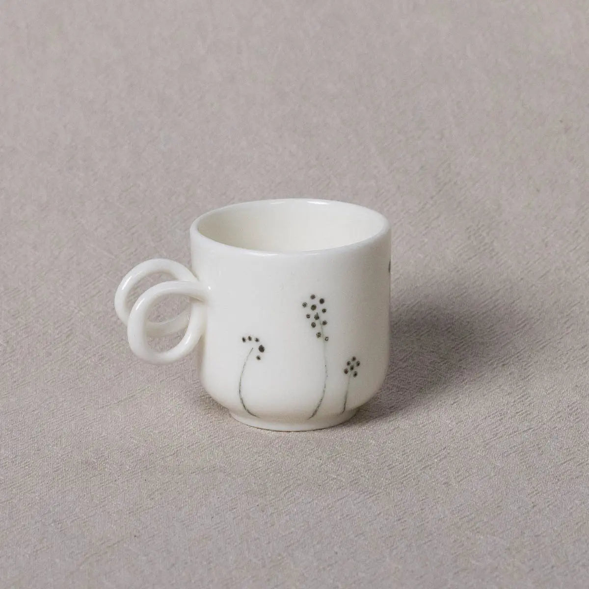 The Espresso Mini Cup, Meadow Flowers by Urchic Porcelain is a small white ceramic espresso cup featuring twisted handles and a minimalist black flower design, meticulously hand-painted to perfection, displayed on a gray fabric background.