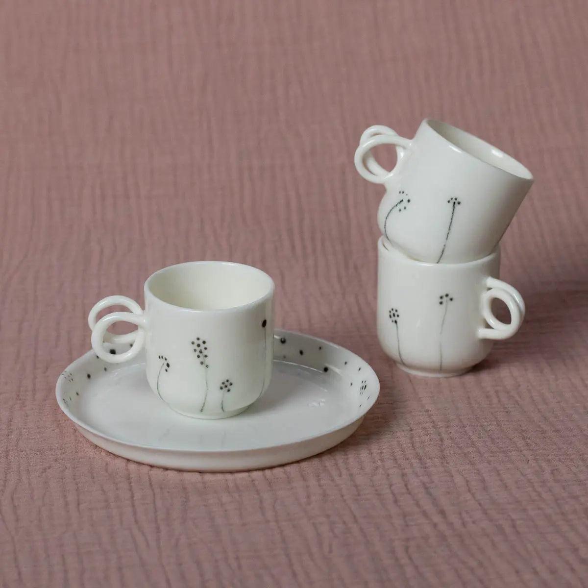 A white ceramic teacup and saucer featuring a minimalist dandelion design and twisted handle, along with two stacked Espresso Mini Cups from Urchic Porcelain's Meadow Flowers collection, are displayed on a pink textured background.