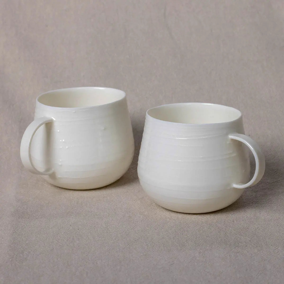 Two identical Nova Cups by Urchic Porcelain, featuring smooth 3D lines and a sleek white finish, sit on a beige fabric surface—perfect for savoring your morning herbal tea.