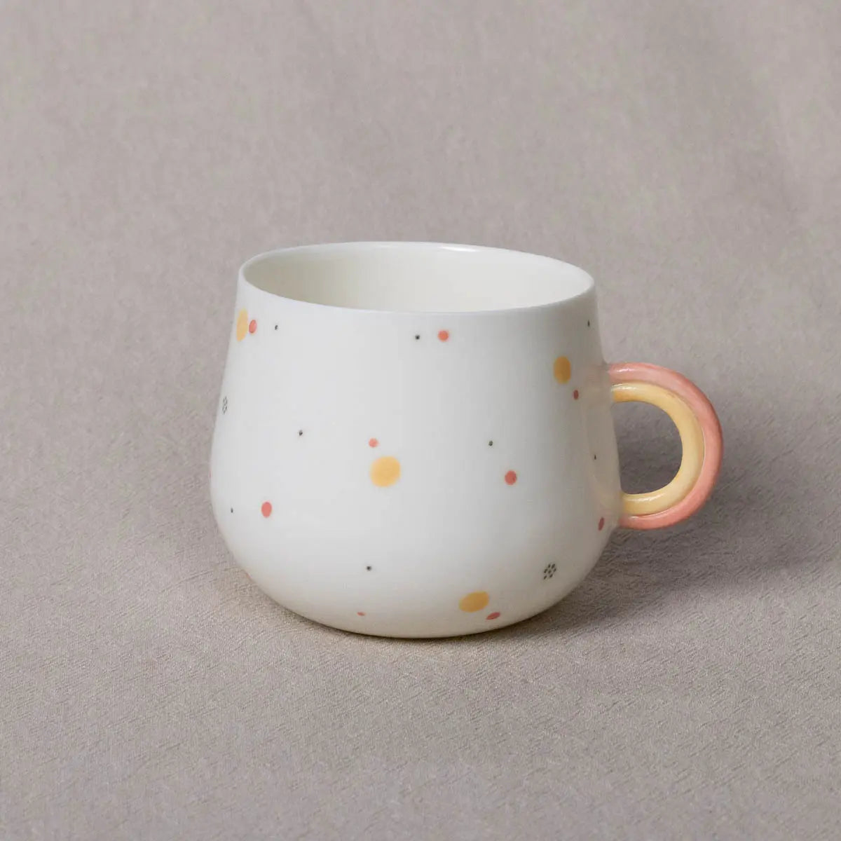 The Nova Cup from Urchic Porcelain is a white, rounded ceramic mug with a rainbow handle accented in pink and yellow. It features small, scattered orange, yellow, and gray dots across its 400 ml surface and is placed on a light gray background.