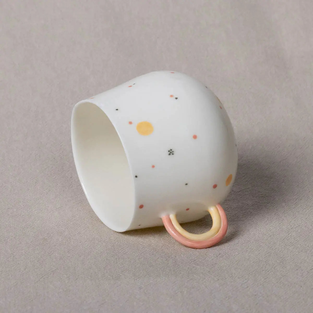 A Nova Cup, rainbow by Urchic Porcelain—a 400 ml white porcelain cup adorned with multi-colored dots and a pastel pink and yellow handle—is lying on its side on a beige surface.