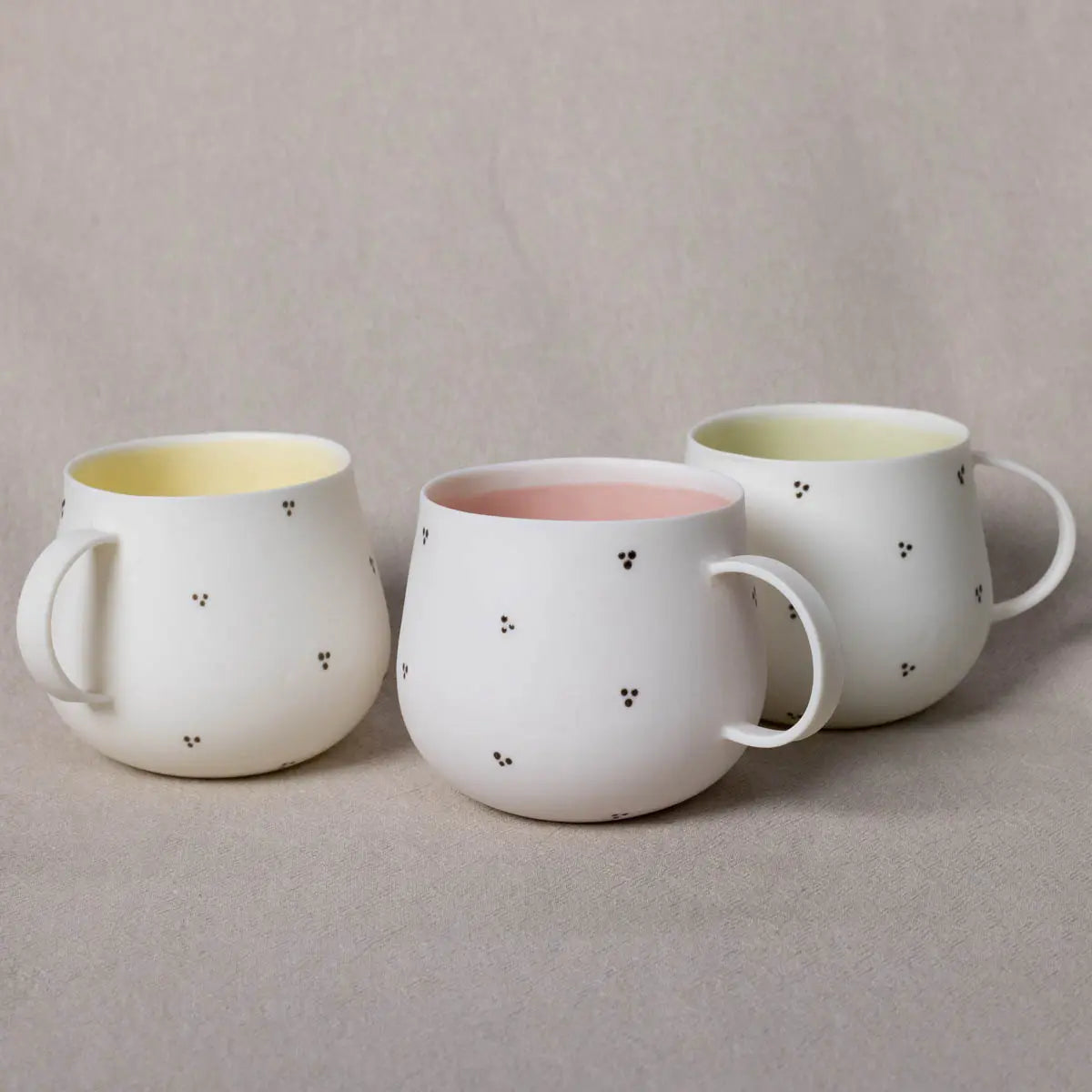 Three Nova Cup, dotty blossoms from Urchic Porcelain, each with large capacities and available in three colors - yellow, pink, and white interiors with decorative patterns - are displayed on a beige surface. The white porcelain cups feature small black dot accents.