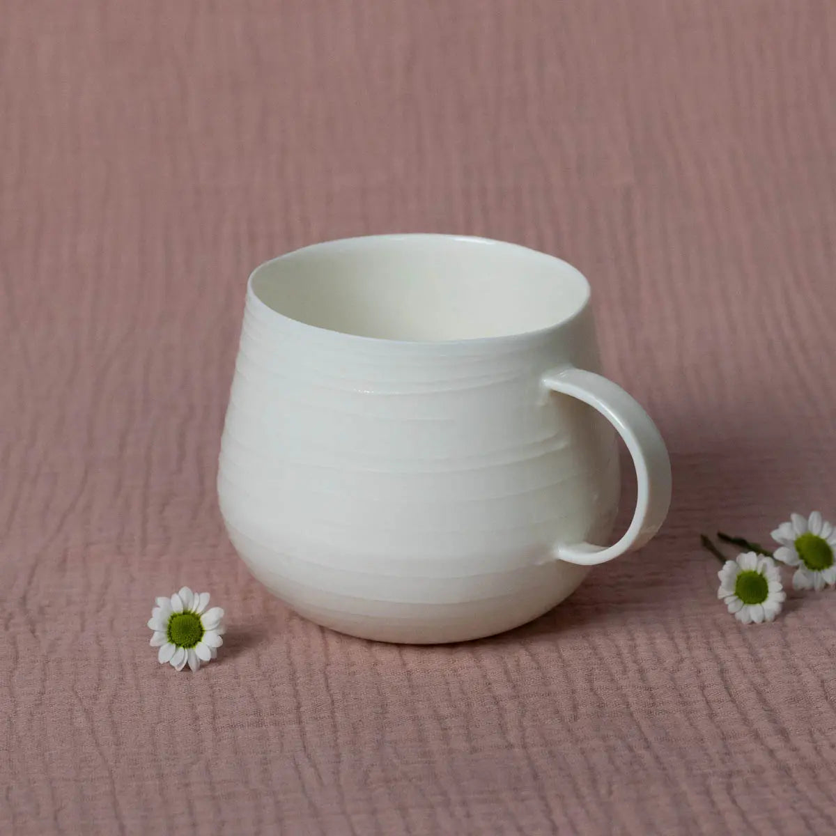 A Nova Cup, featuring smooth 3D lines in white porcelain from Urchic Porcelain, is placed on a textured surface beside two small white daisy flowers, perfect for your morning herbal tea.