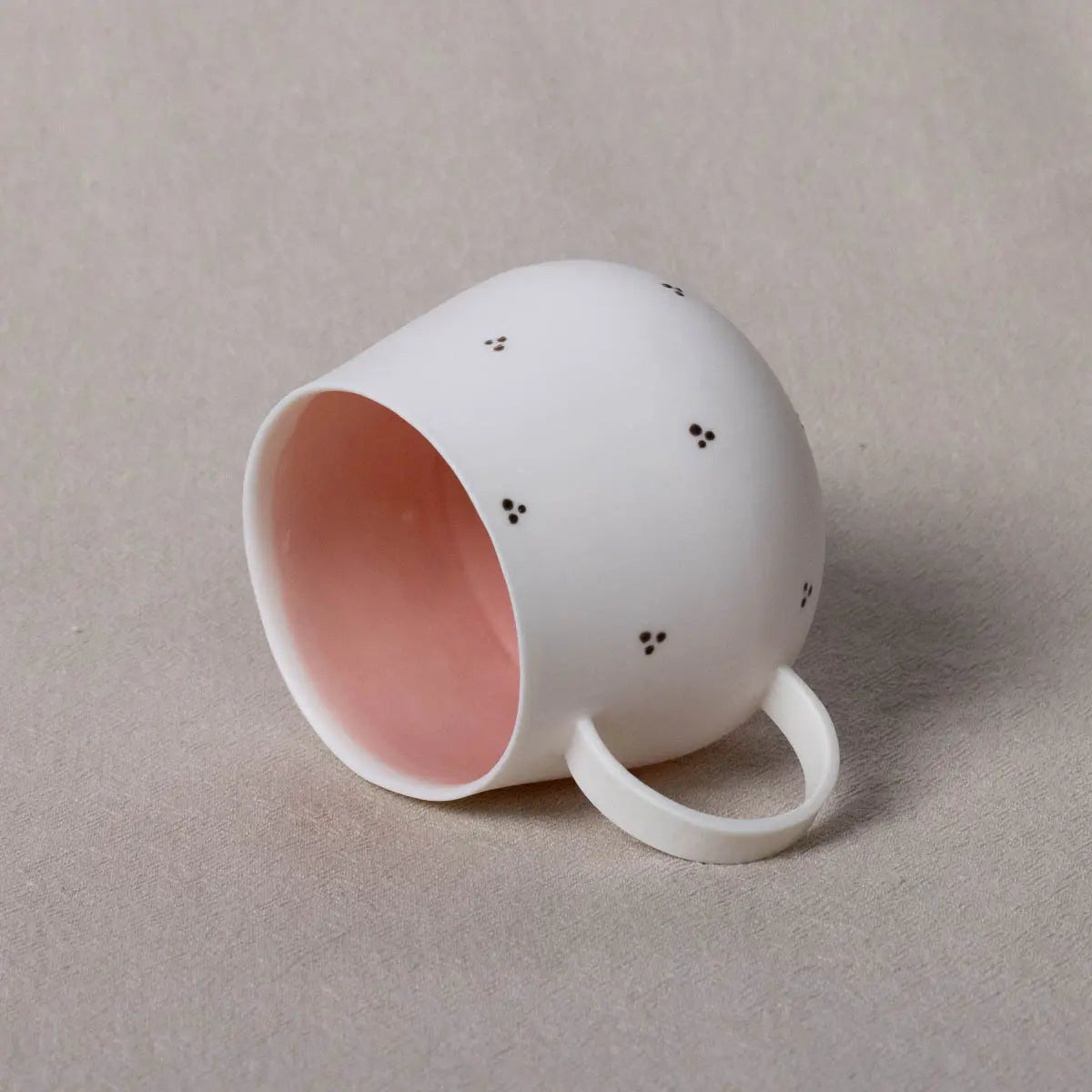 The Nova Cup by Urchic Porcelain, featuring a dotty blossoms design in white with small black polka dots on the outside and a soft pink interior, is placed on its side on a beige surface. This charming decorative pattern enhances its spacious capacity.