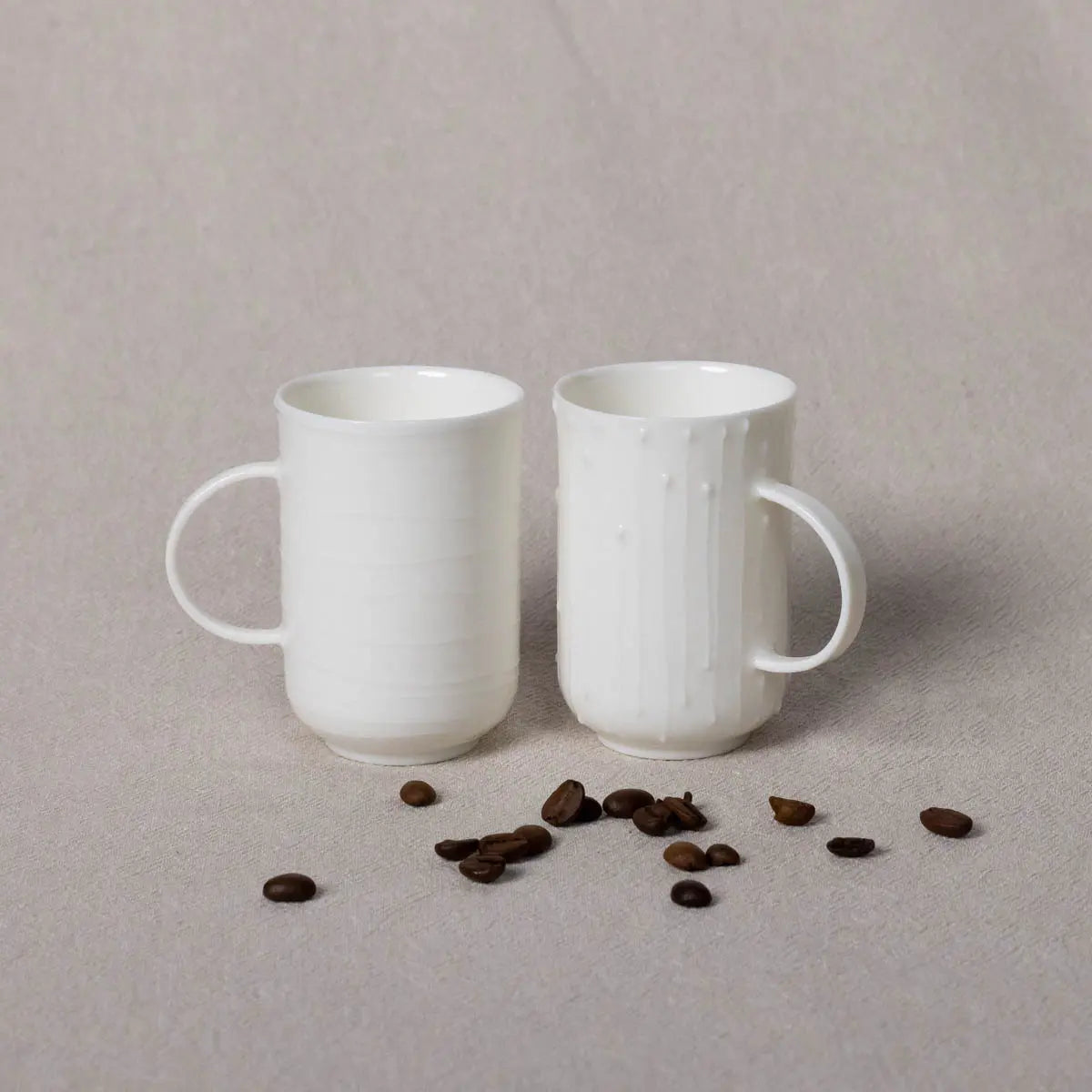 Two Soulmates porcelain coffee cups with a Lines pattern from Urchic Porcelain are placed next to each other on a beige surface, complemented by scattered coffee beans in front of them, creating an inviting coffee set.