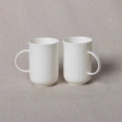 Two white porcelain mugs with handles, placed side by side on a neutral-colored surface. The mug on the left has a smooth finish, while the one on the right features a subtle textured design. This Soulmates Porcelain Coffee Set of Two Cups from Urchic Porcelain captures the essence of handmade craftsmanship.