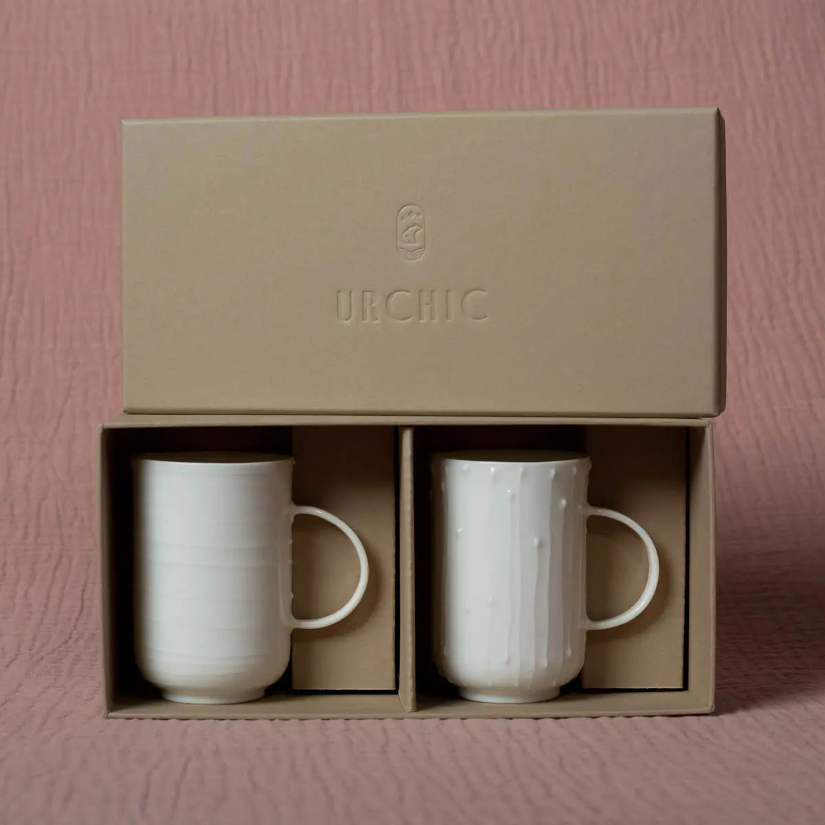 Two white porcelain cups with a stylish lines pattern sit in individual slots of a beige cardboard box adorned with "Urchic Porcelain" on the top. The background features a textured pink surface, creating an ideal setting to showcase this elegant "Soulmates" coffee set of two cups.