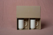 Two white handmade porcelain mugs from the "Soulmates" coffee set are displayed in a beige box with two compartments. "URCHIC" is embossed on the front of the box, showcasing this elegant set by Urchic Porcelain. The background is pink with a textured pattern.