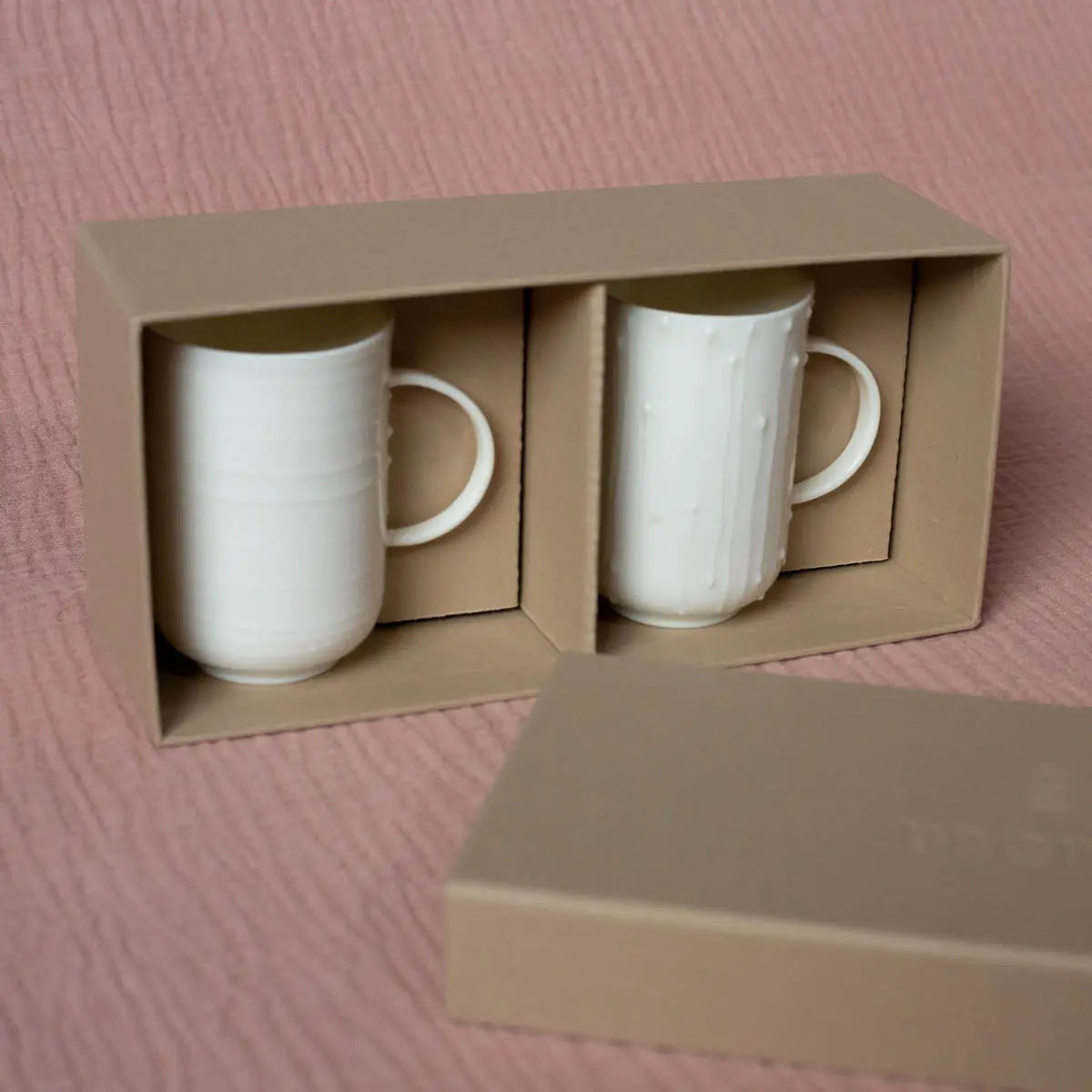 A set of two handmade Soulmates porcelain cups with a Lines pattern from Urchic Porcelain, placed in a beige cardboard box with a lid nearby on a pink textured surface.