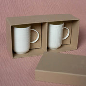 A set of two handmade Soulmates porcelain cups with a Lines pattern from Urchic Porcelain, placed in a beige cardboard box with a lid nearby on a pink textured surface.