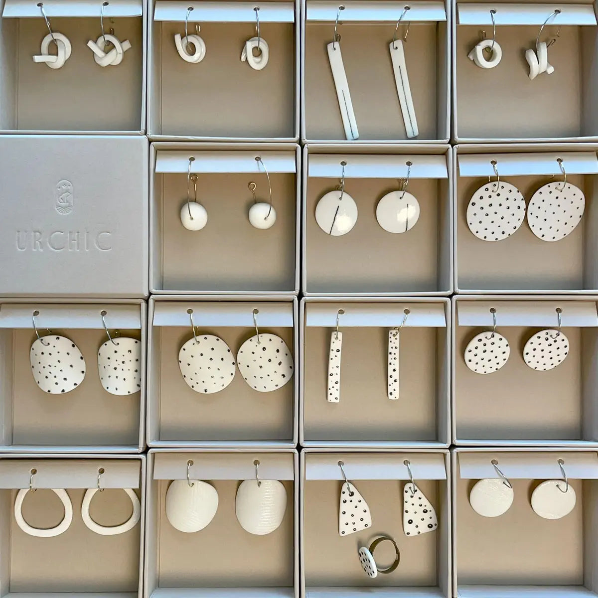 Eighteen pairs of Urchic Porcelain Earrings - Polka dots, each handcrafted with various shapes and patterns including dots and stripes, are mounted on a display board. A box labeled "Urchic Porcelain" is included on the left side of the display.